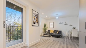 Ein modernes Wohnzimmer mit grauem Sofa, einem Couchtisch und einem Essbereich mit weißen Stühlen. Ein Balkon mit Metallgeländer bietet Blick auf Bäume.