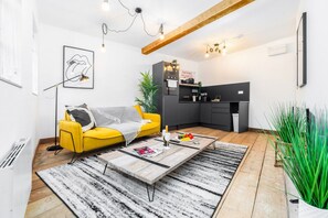 A modern living room with a yellow sofa, a wooden coffee table, and a kitchen area with a sign prohibiting smoking.