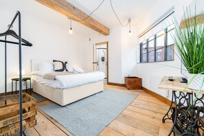 A bedroom with a bed, a wooden floor, a grey rug, a wooden table, and a hanging plant.