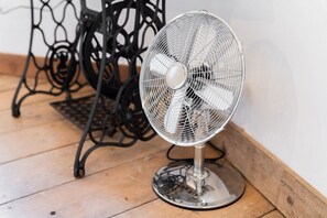 A standing fan with a metal base and a protective grill, positioned on a wooden floor near a black metal table.