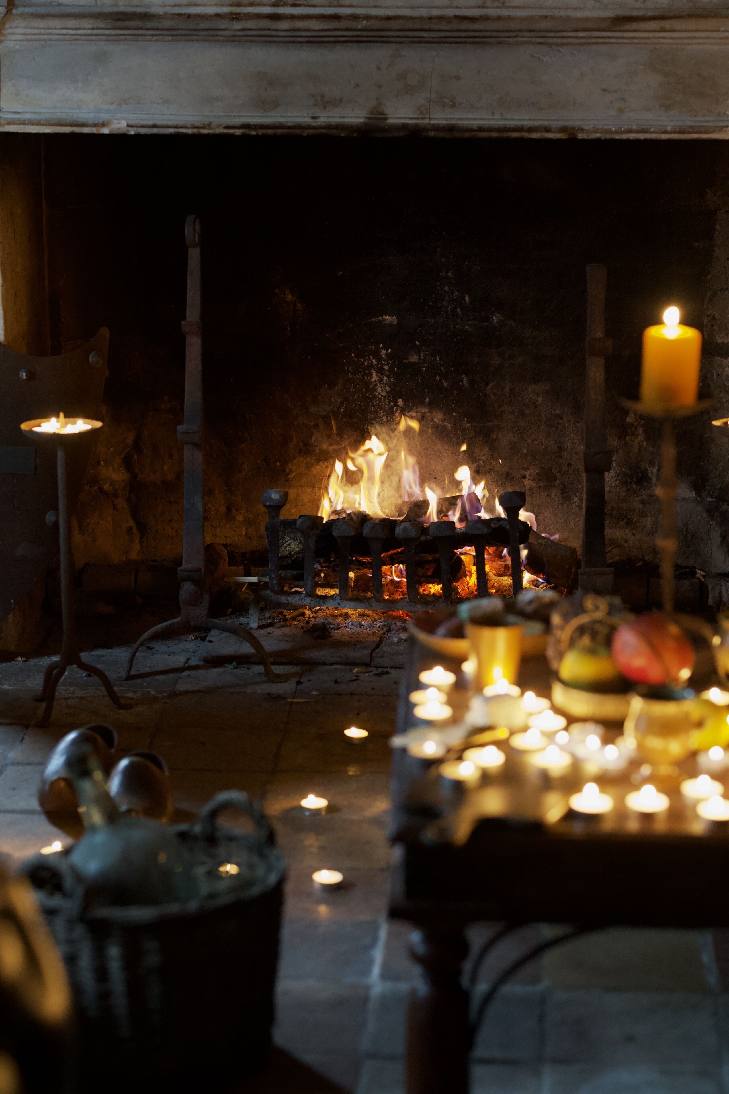 A traditional fireplace with a fire, surrounded by candles and a table with fruits and a candle holder.