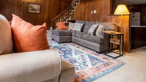 A cozy living room with a sofa, patterned rug, and a chandelier. A staircase leads to an upper level with a kitchen area visible. - Spacious Chalet, Skiing 5 Minutes to Jay Peak, Vermont! (Montgomery)