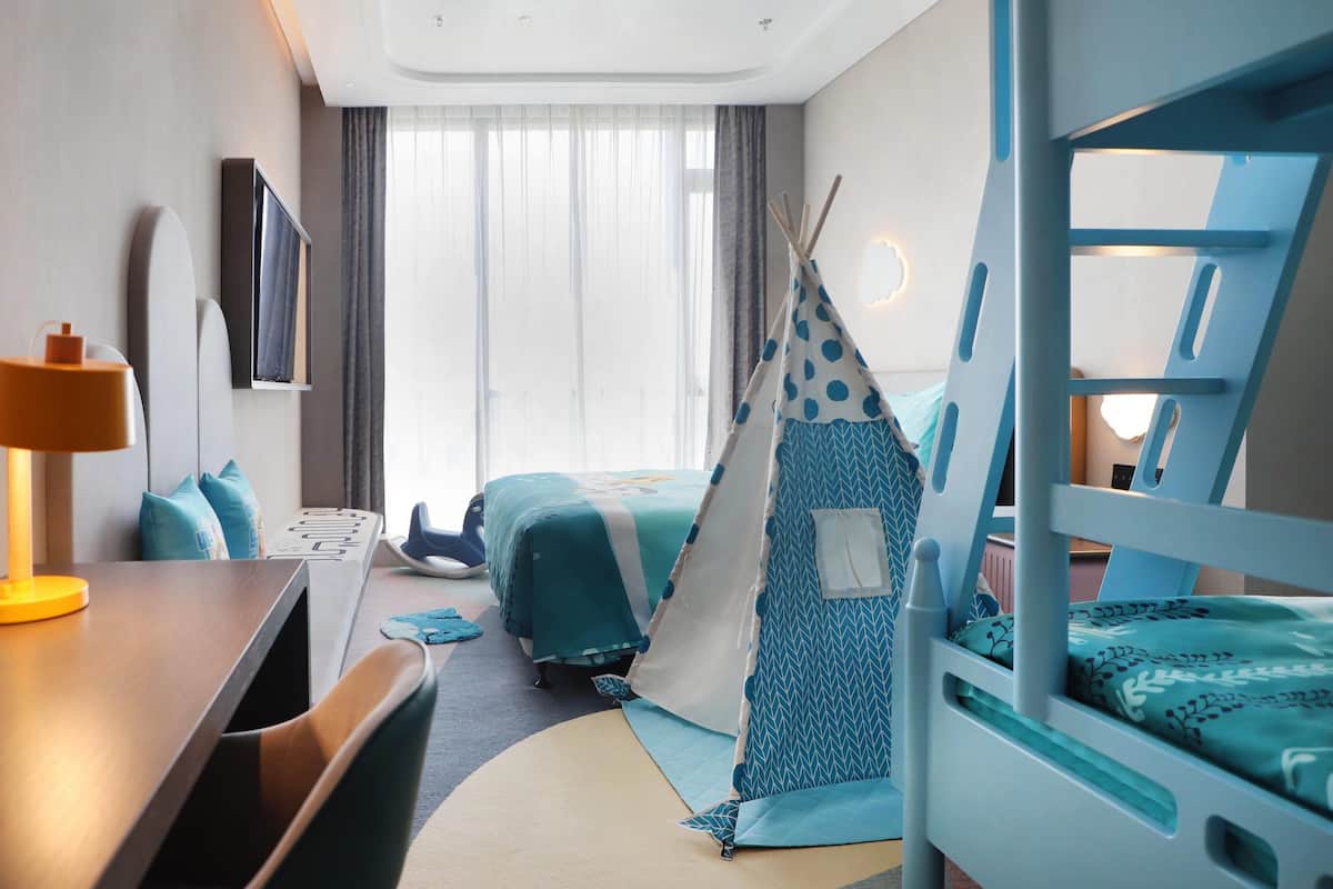 Une chambre d’hôtel avec un lit superposé, un bureau avec une lampe, un lit recouvert d’une courtepointe à pois bleus et blancs, une tente de style tipi et une grande fenêtre avec des rideaux légers.