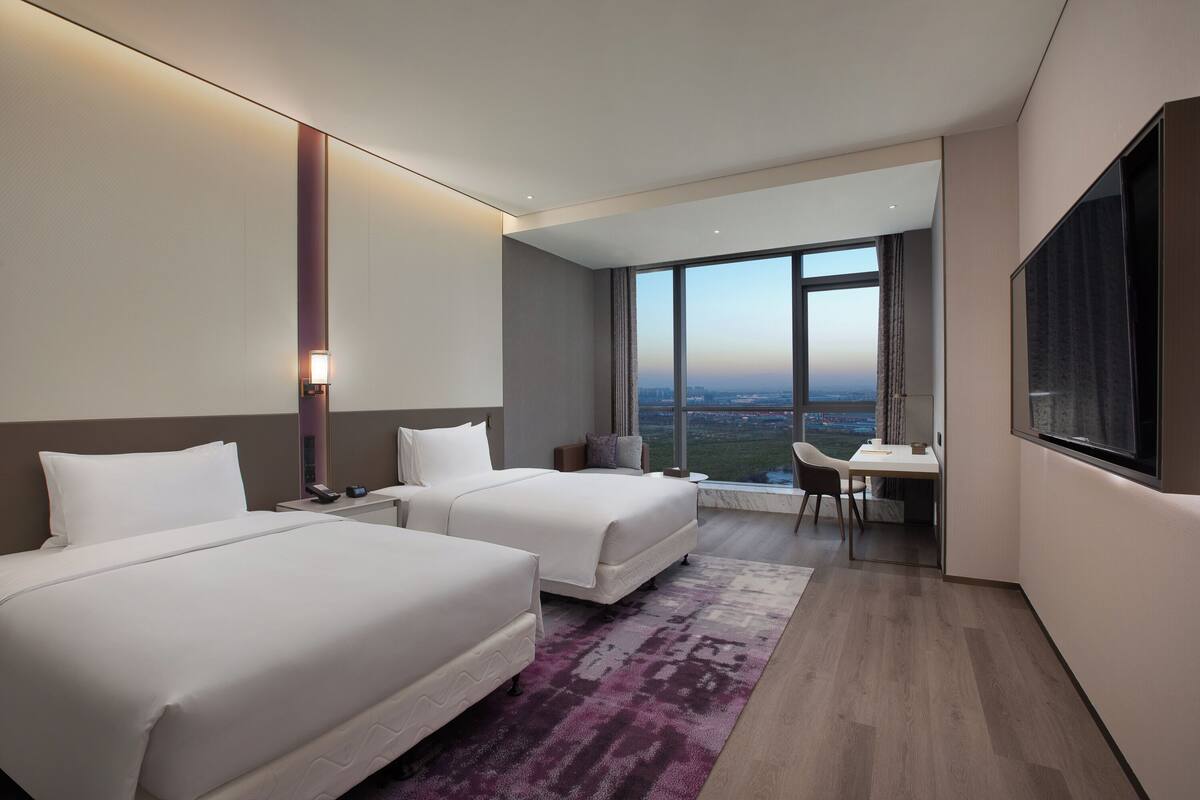 Une chambre d’hôtel moderne avec deux lits, un bureau et une grande fenêtre donnant sur la ville.