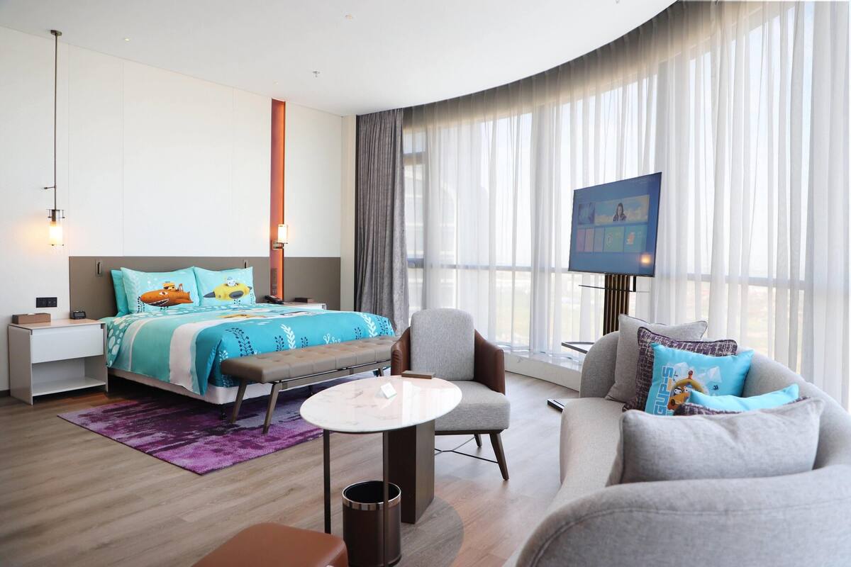 Une chambre d’hôtel moderne équipée d’un lit, d’un canapé, d’une petite table, d’une télévision et d’une grande fenêtre avec des rideaux.