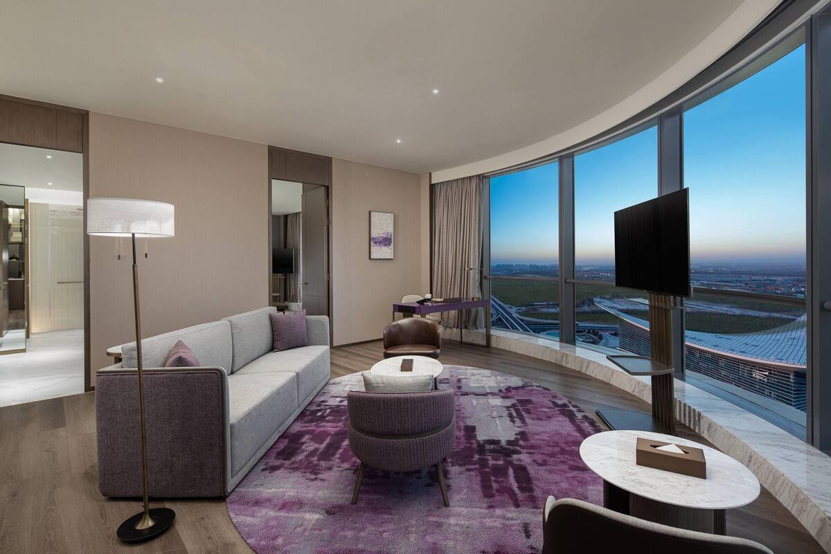 Une chambre d’hôtel moderne avec une grande fenêtre, un canapé, une table ronde et un lampadaire.