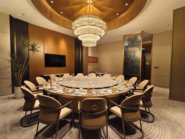 Un salón de conferencias con una mesa redonda, sillas color beige, un candelabro, una planta en maceta y un cuadro grande en la pared.