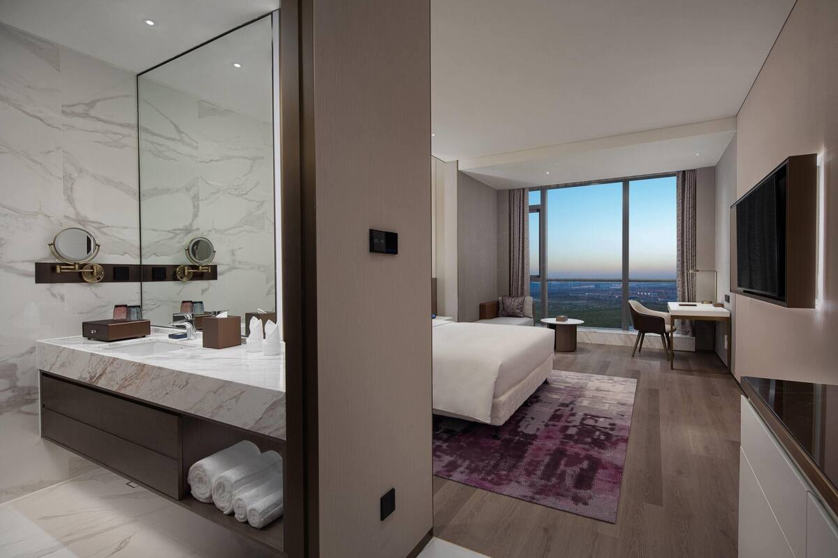 Une chambre d’hôtel moderne dotée d’un grand lit, d’un bureau, d’une télévision et d’une salle de bain avec un grand miroir et un comptoir en marbre.