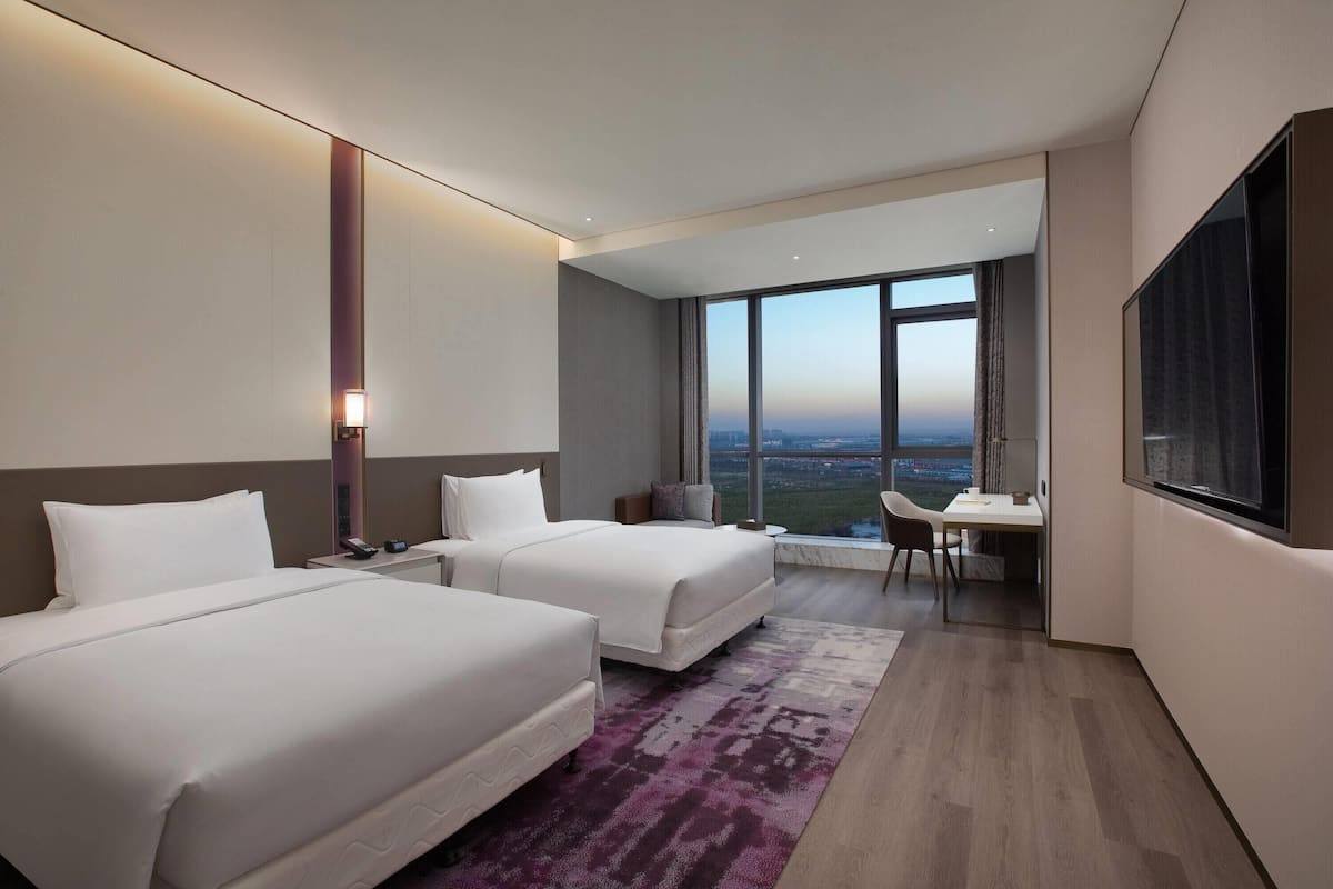 A hotel room with two beds, a desk, a chair, a large window with a city view, and a flat-screen TV.