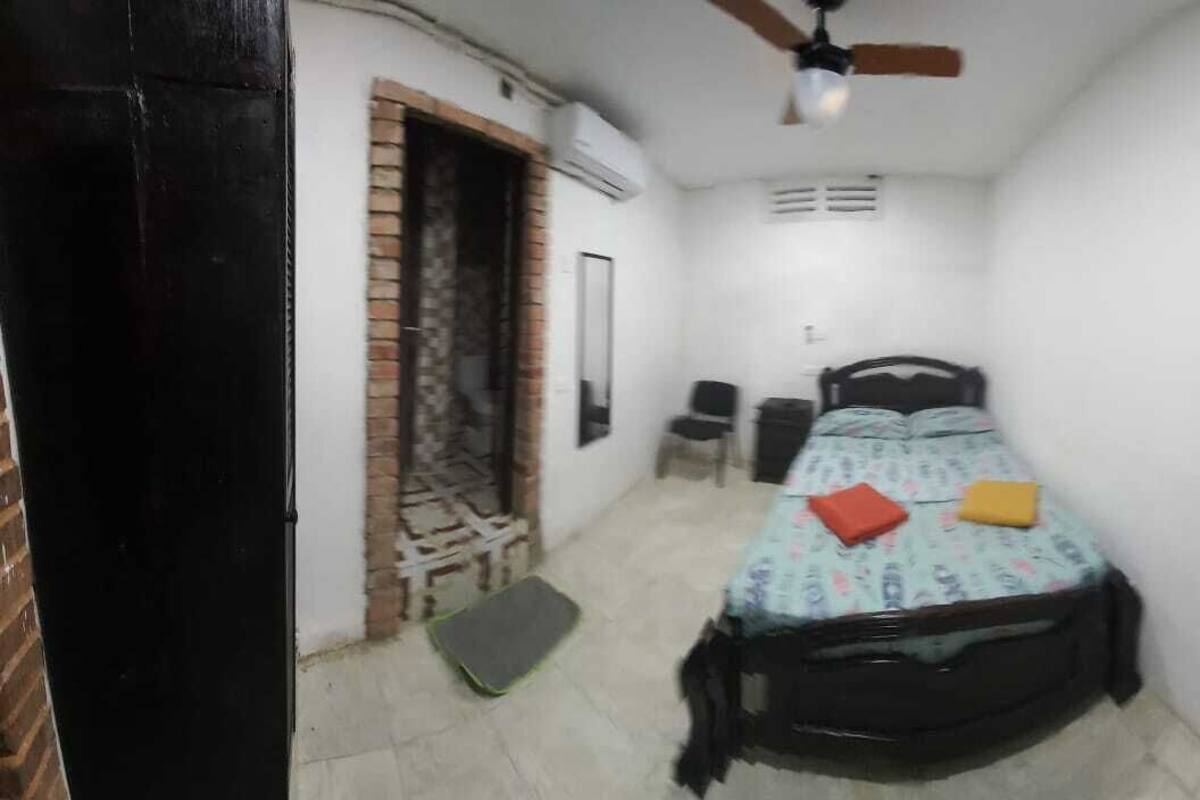A small, narrow room with a single bed, a ceiling fan, and a door leading to another room.