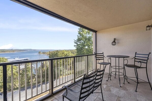 A balcony with a view of a lake, a table, two chairs, and a small lamp.