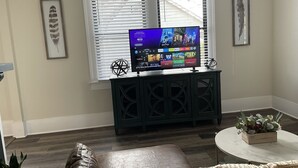 Een woonkamer met een flatscreen televisie op een donker houten standaard, een leren bank, een ronde salontafel met een plant en twee ramen met jaloezieën.