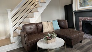 Een moderne woonkamer met een bruine leren modulaire bank, een ronde salontafel en een open haard. Een trap met houten treden en een witte balustrade is op de achtergrond zichtbaar.