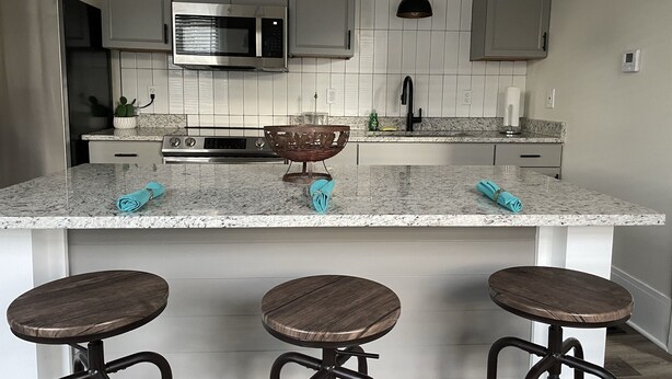 Een moderne keuken met een centraal eiland, barstoelen, roestvrijstalen apparatuur en hangende verlichting.