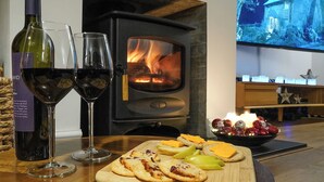 Une pièce chaleureuse avec une cheminée, un téléviseur diffusant une image d’une maison, et une table avec des collations et des verres à vin.