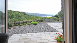 La vue depuis une pièce dotée d’une grande fenêtre donnant sur un jardin avec un mur de pierre, un chemin de gravier et un paysage lointain.