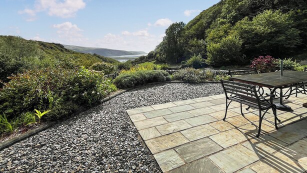 Une terrasse de jardin avec un banc, entourée de gravier et de dalles de pierre, offrant une vue sur une colline verdoyante et sur la mer au loin.