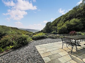 Eine Gartenpatio mit einer Bank, umgeben von Kies und Steinplatten, mit Blick auf einen grünen Hang und eine ferne Meerblick.