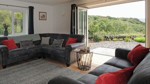 Un salon moderne avec un canapé d’angle, une petite table ronde et une grande fenêtre donnant sur un paysage verdoyant et vallonné.