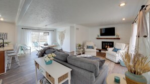 Ein geräumiges Wohnzimmer mit grauem Sofa, weißen Armstühlen, einem flachbildfernseher über einem Kamin und einem Essbereich mit weißen Stühlen und einem Tisch.