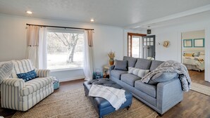 Ein Wohnzimmer mit einem gestreiften Sessel, einem grauen Sofa und einem blauen Hocker. Es gibt ein Fenster mit Vorhängen, eine Vase mit Trockenblumen und eine Tür, an der ein Hut hängt.