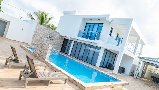 Un área moderna junto a la piscina con una pileta rectangular, sillones de madera para tomar el sol y un edificio de dos pisos con ventanas amplias y un balcón.