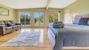 A bedroom with a large bed, a sofa, and a view of trees through large windows. - Angel Bear Chalet- Private Lake/Beach/Sauna/Hot Tub/Gym/Firepit/Float Trampoline (Peru)