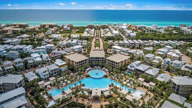 An aerial view of a resort with multiple buildings, a central swimming pool, and a view of the ocean.
