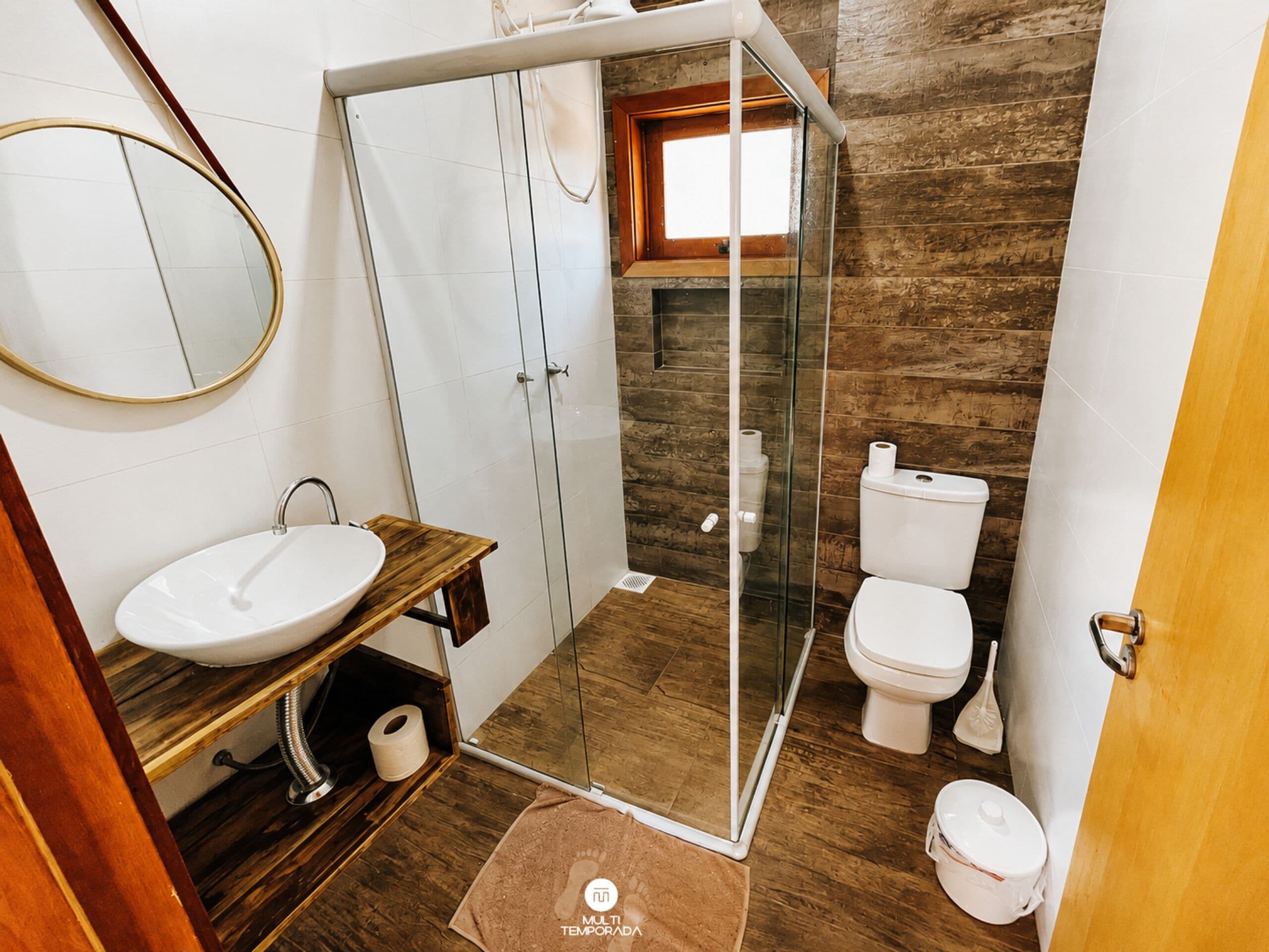Un baño con ducha de vidrio, tocador de madera, lavamanos blanco y un inodoro.