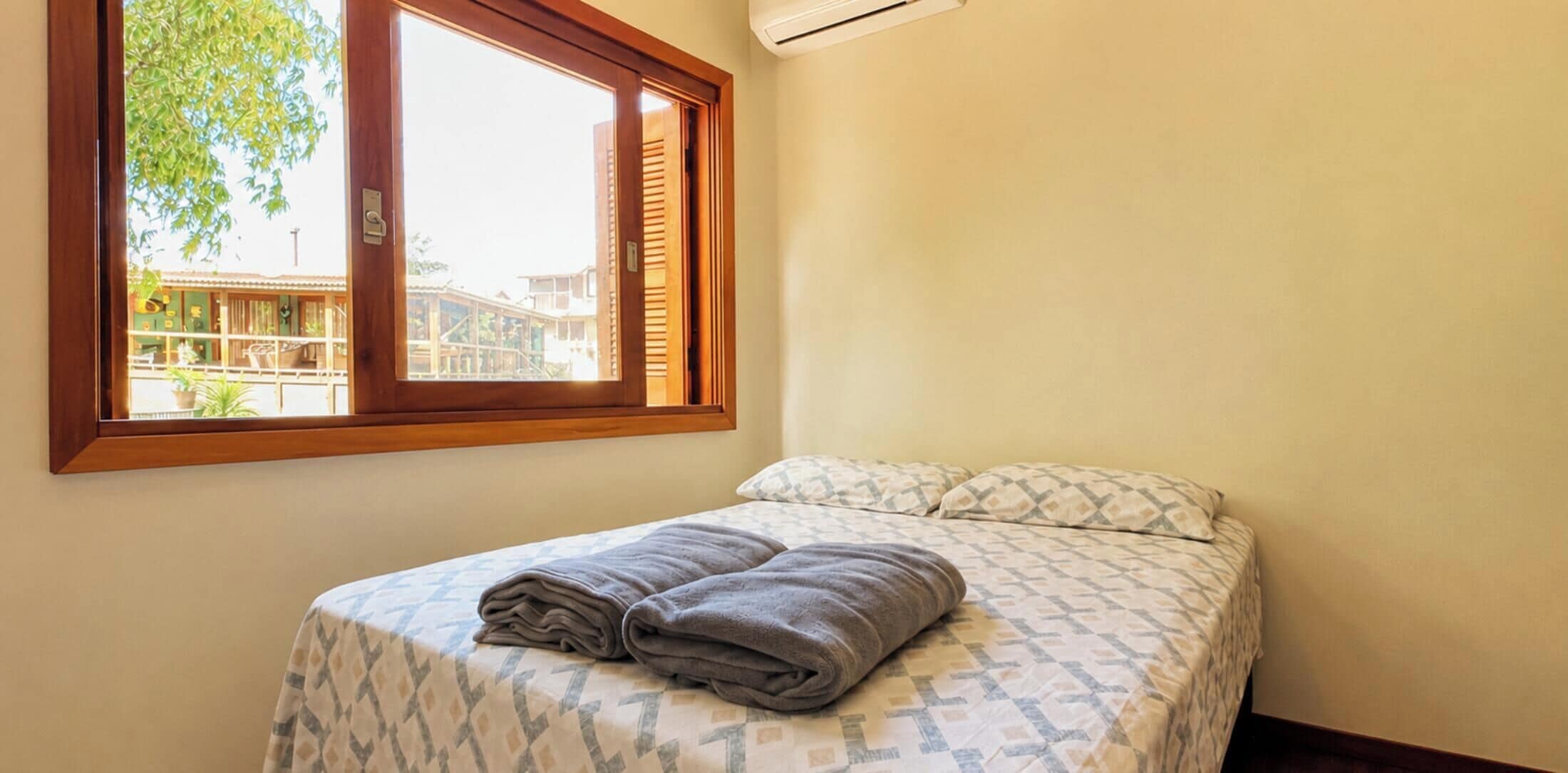 A single bed with a patterned bedspread and two pillows, a window with blinds, and an air conditioning unit on the wall.