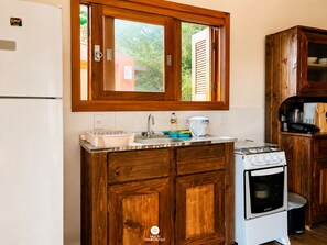 A kitchen with wooden cabinets, a white refrigerator, a stove, and a window with blinds. - Rosa Pantai Beach Village (Imbituba)