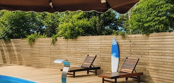 A poolside area with wooden loungers and a surfboard leaning against a wooden fence.