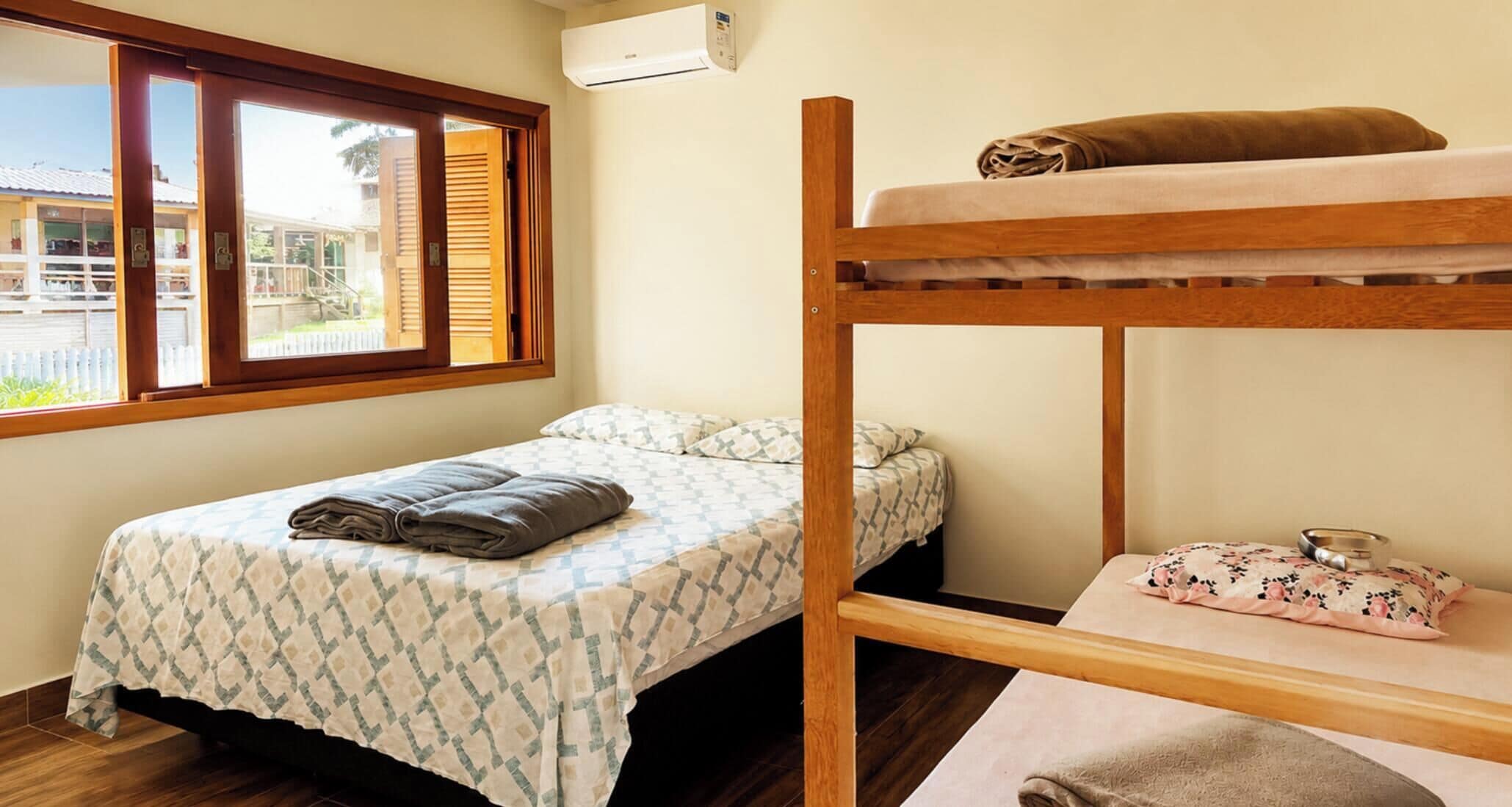 A room with a bunk bed, a single bed with patterned bedding, a window with blinds, and an air conditioning unit.
