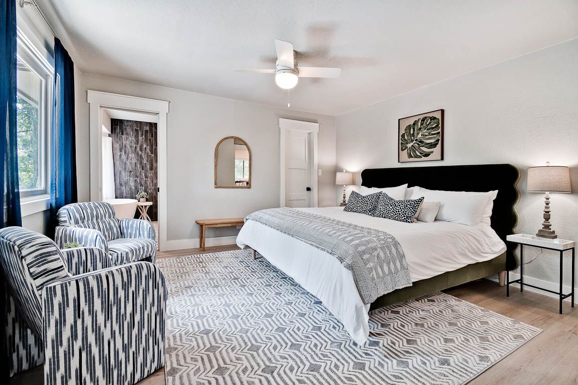 A bedroom with a bed, two armchairs, a bedside table, a floor lamp, a ceiling fan, and a patterned rug.