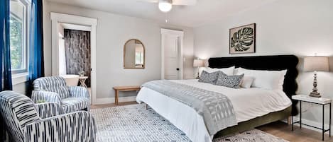 A bedroom with a bed, two armchairs, a bedside table, a floor lamp, a ceiling fan, and a patterned rug.