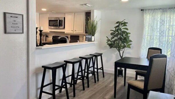 A modern kitchen with white cabinets, a microwave, and a coffee machine. There are four black stools and a table with chairs. A potted plant is on the counter, and a framed welcome sign is on the wall. - Close to Disney and the Beaches Full House 3/3 (Orlando)