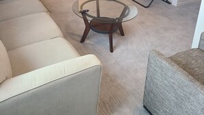 Una sala de estar con una mesa de centro de vidrio, un sofá, una silla negra, un televisor montado en la pared y una zona de cocina al fondo.