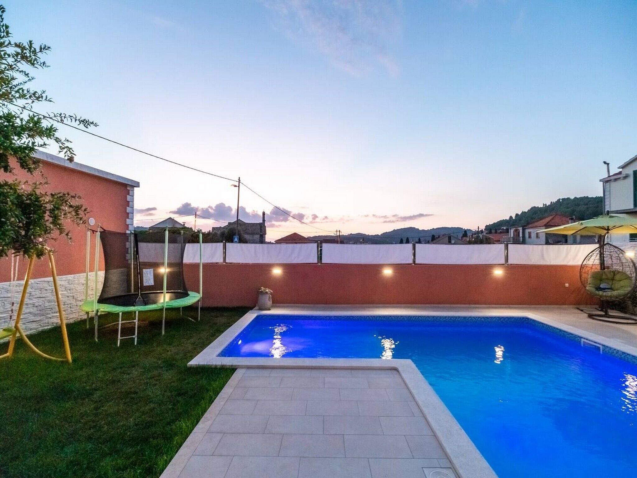 Una piscina rectangular con una solana empavedada, rodeada por un muro de ladrillo bajo y un área verde.