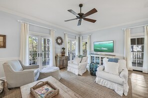 Relax in this spacious living room with a high ceiling, ceiling fan, and large window offering a view of lush greenery.
