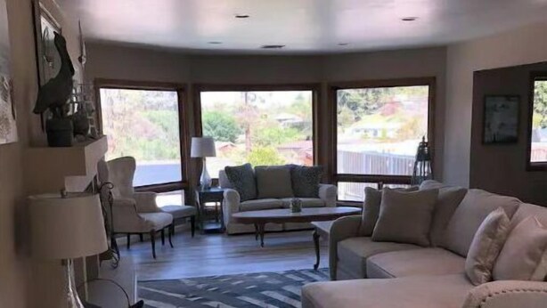 A spacious living room with a beige sofa, a coffee table, and a patterned rug. There are large windows providing a view of the outdoors. - Beautiful 3,000 Sq Ft Tropical Oasis (El Cajon)