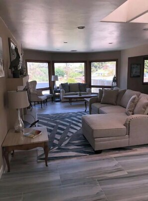 A spacious living room with a beige sofa, a coffee table, and a patterned rug. There are large windows providing a view of the outdoors. - Beautiful 3,000 Sq Ft Tropical Oasis (El Cajon)