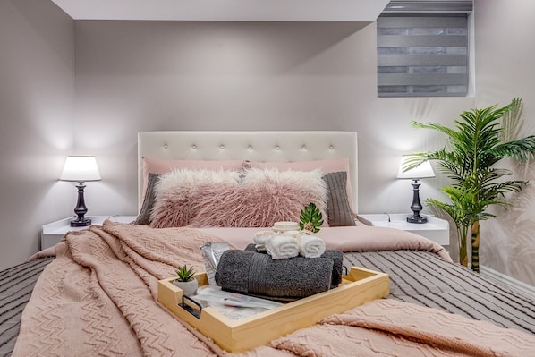 A neatly made bed with a fluffy pink pillow, a bedside table with a lamp, and a tray with towels and a small plant.