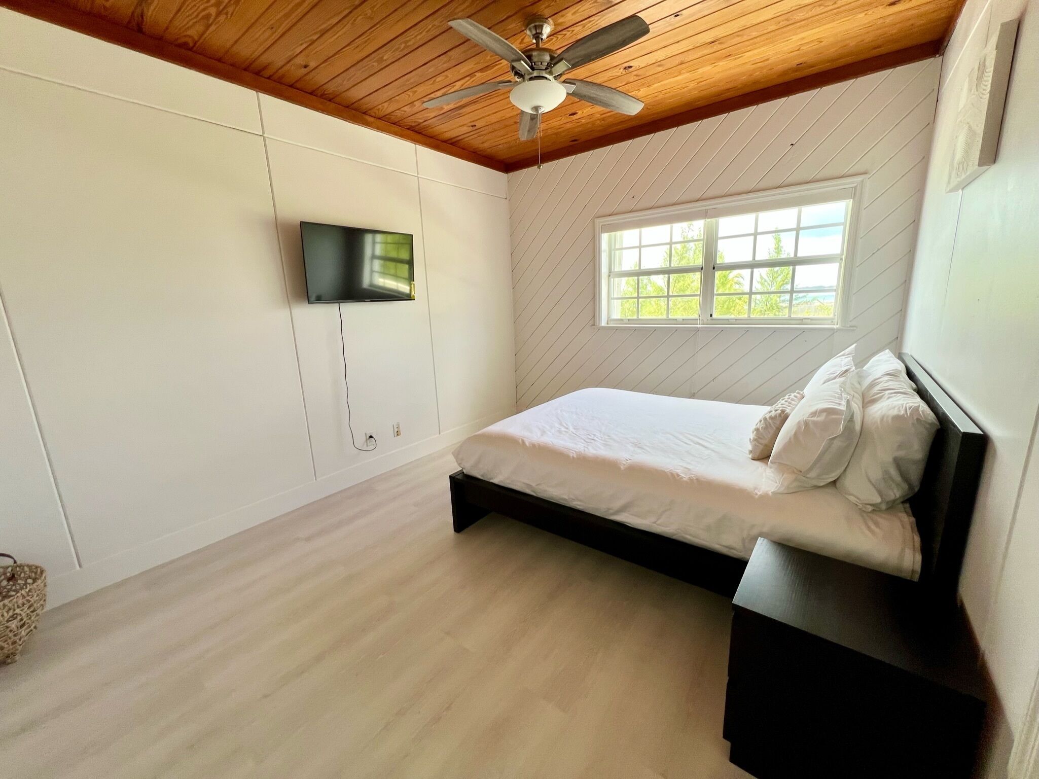 A bedroom with a bed, a ceiling fan, a TV mounted on the wall, and a window with a view of greenery.