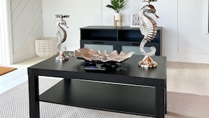 A modern living room with a black coffee table, two decorative seahorse sculptures, a woven basket, and a potted plant.