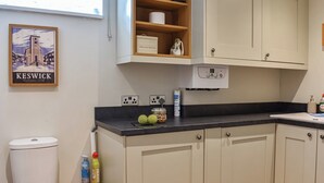Une cuisine avec un plan de travail noir, des armoires blanches et une étagère en bois. Un tableau encadré est accroché au mur, et il y a une fenêtre avec des stores.