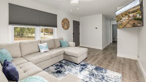 A modern living room with a sectional sofa, a ceiling fan, a large window with blinds, and a flat-screen TV mounted on the wall. - Eudora House with Epic Game Room (Eudora)