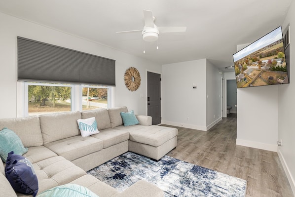 A modern living room with a sectional sofa, a ceiling fan, a large window with blinds, and a flat-screen TV mounted on the wall.