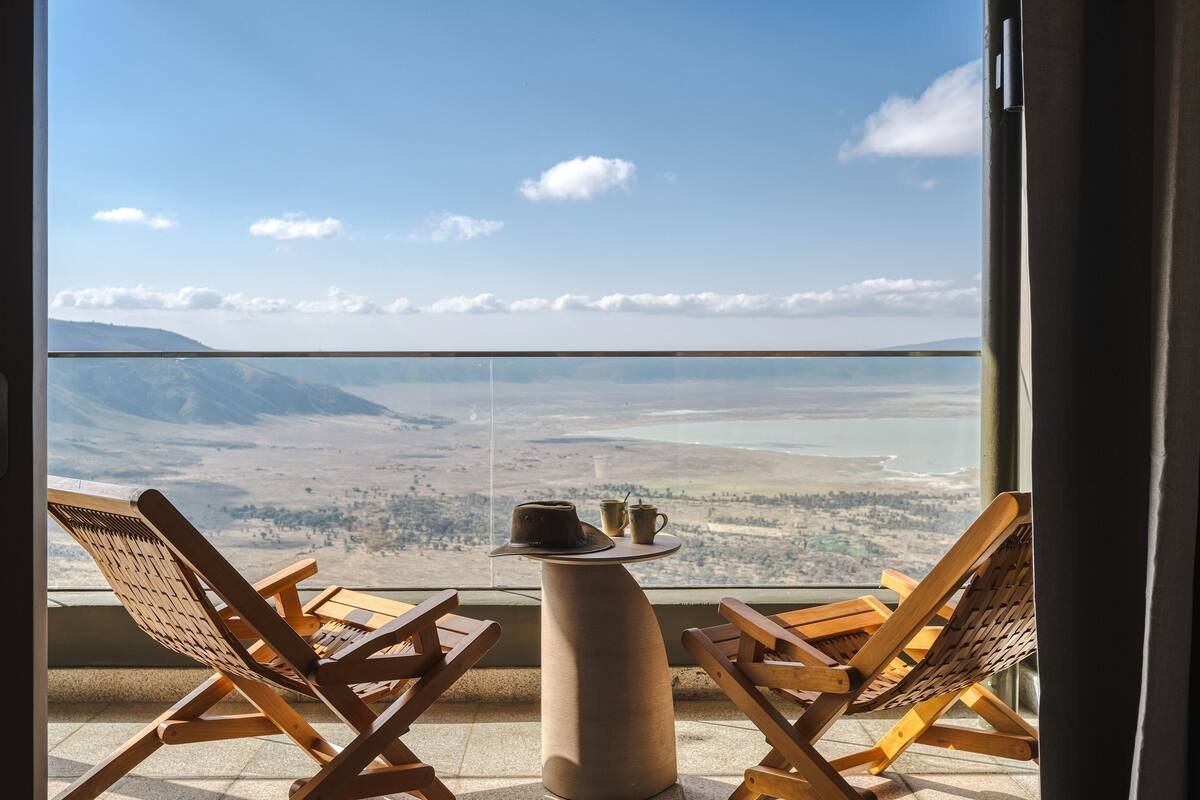 A balcony with two wooden lounge chairs and a small table, overlooking a scenic landscape with mountains and a body of water.