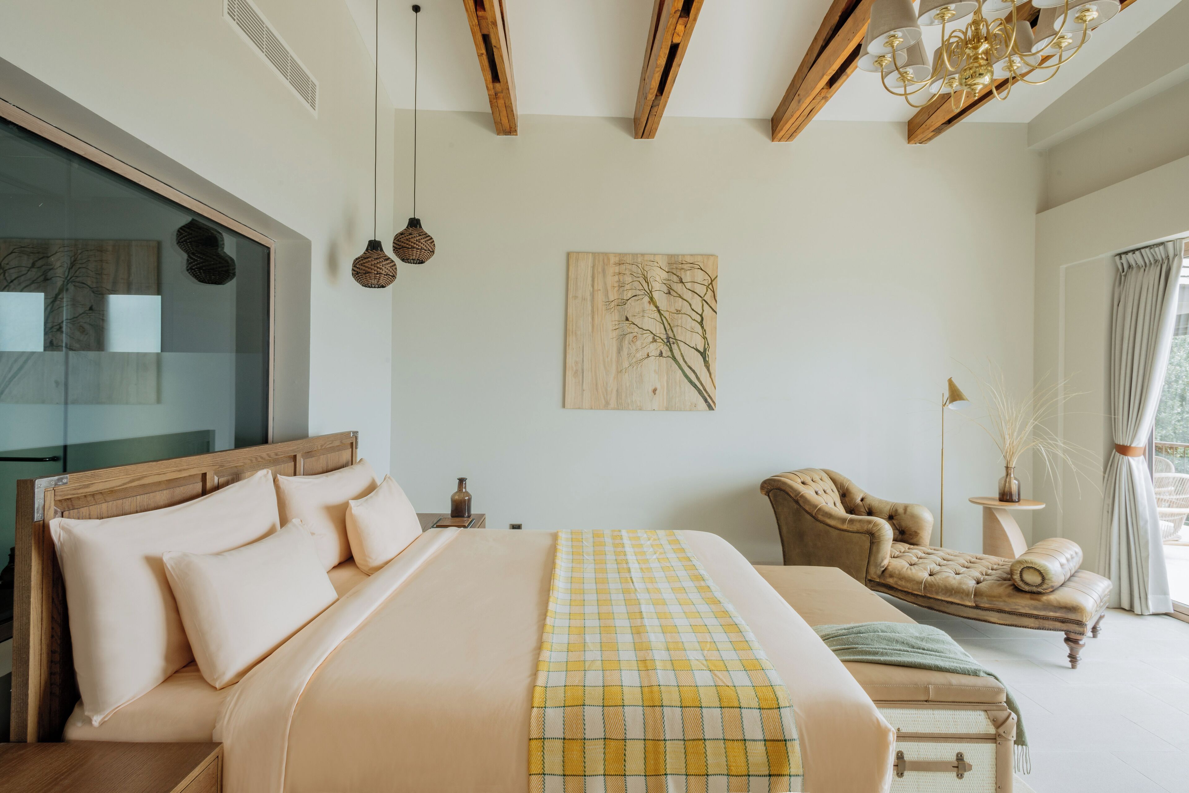 A bedroom with a large bed, a wooden headboard, a chandelier, a painting, a window with curtains, and a side table with a vase.