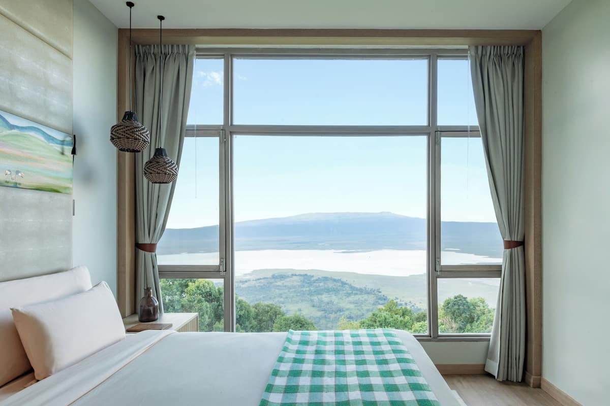 A bedroom with a large window, a bed with white linens, a green checkered throw, and a painting on the wall.